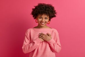 Lovely curly young woman presses palms to heart, feels touched and grateful, looks with admiration at something, keeps all good memories, dressed in pink jumper, isolated over rosy background